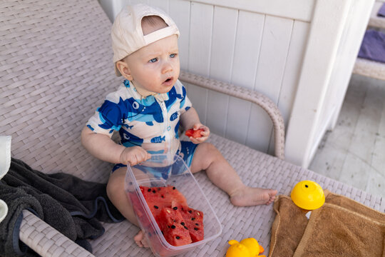 Baby in swimwear enjoys watermelon slices while sitting on a lounge chair