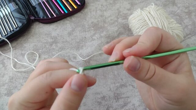 Hands crocheting a basic yarn chain using a green aluminum hook and white yarn. Horizontal stock footage for craft tutorials, DIY knitting, handmade hobbies, and creative learning vlogs.