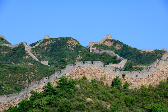 The Jinshanling Great Wall, an ancient stone fortification, winds across lush green mountain slopes under a vivid blue sky, with steep stone steps and aged watchtowers rising amid dense vegetation.