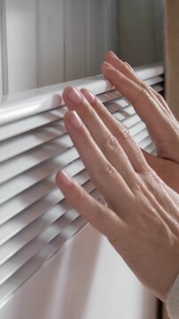 Close up of female palms touching electric convector heater. Vertical video. Woman in knitted sweater warming hands in cold room. Energy crisis, high utility bills. Cold home, winter poverty.