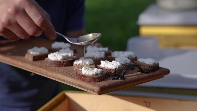 Hand drizzling golden honey from a spoon onto small rye bread slices topped with cottage cheese, arranged on a wooden serving tray in warm sunlight