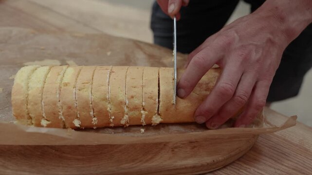 Close-up of hands cutting a homemade sponge roll cake into even slices on parchment paper over a wooden serving board, with cream filling visible between layers