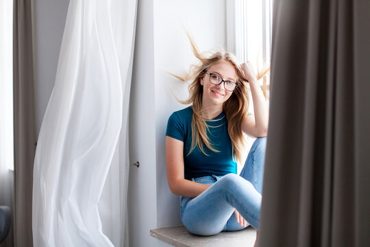 Relaxed young woman sitting on windowsill at home. Happy girl enjoying fresh air and wind by window. Concept of female mental health, slow living, social rest, mindfulness. Copy space