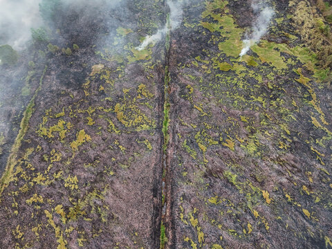 Aerial view of a rugged volcanic lava field with rising smoke plumes and patches of green vegetation in Indonesia.
