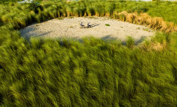 Aerial view of two people riding bicycles on a sandy patch surrounded by tall green grass in Dhaka, Dhaka Division, Bangladesh.