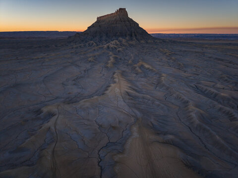 Aerial view of Factory Butte rising above the rugged, eroded badlands of the desert landscape under a soft golden hour sky in Wayne County, Utah, United States.
