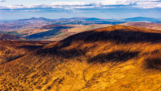 Wind turbines are visible in the distance on a hill crest against a backdrop of blue sky and clouds.