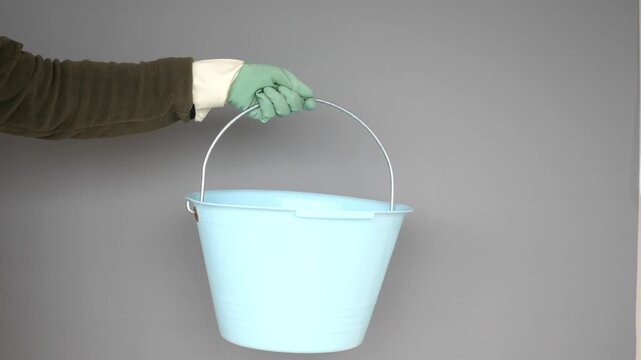 Close up of person wearing green work gloves and holding blue plastic bucket against grey background
