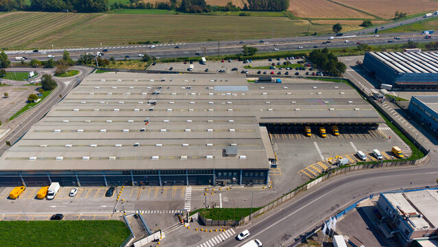 Aerial view of the Esselunga Spa logistics facility with a large warehouse, loading docks, yellow delivery trucks, and parking areas in Sesto Ulteriano, Lombardia, Italy.