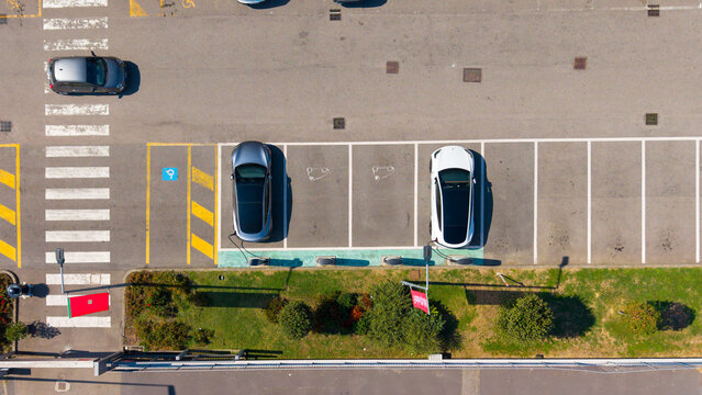 Aerial view of a Tesla Supercharger EV station with two electric vehicles parked and charging in a marked lot beside a shopping center in San Giuliano Milanese, Lombardy, Italy.