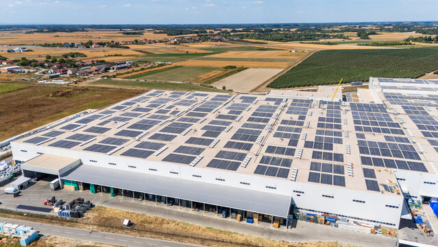 Aerial view of a large industrial warehouse with extensive rooftop solar panel installation surrounded by agricultural fields and rural landscape in Sabac, Vojvodina, Serbia.
