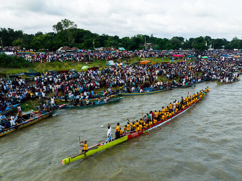 Aerial view of a traditional boat race with rowers in yellow shirts on long narrow boats and large crowds gathered along the riverbank in Manikganj, Dhaka Division, Bangladesh.