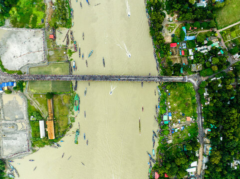 Aerial view of a crowded bridge over a river with numerous small boats navigating the water, surrounded by lush green riverbanks and rural landscape in Manikganj, Dhaka Division, Bangladesh.