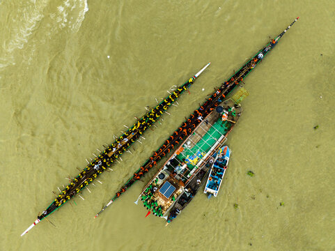 Aerial view of traditional long boats racing on the river with many rowers in colorful uniforms, surrounded by a support boat and spectators in Manikganj, Dhaka Division, Bangladesh.