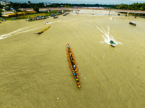 Aerial view of a traditional boat race with rowers in yellow shirts paddling long boats on the river near a bridge in Manikganj, Dhaka Division, Bangladesh.