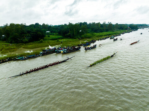 Aerial view of a traditional boat race on a river with long wooden boats filled with rowers and spectators lining the riverbank in Manikganj, Dhaka Division, Bangladesh.