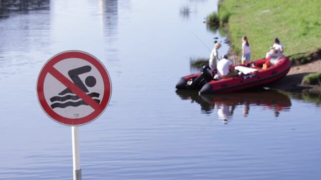 Prominent "no swimming" sign stands in calm waterfront with red prohibition border and black pictogram; in background, people gather near red inflatable boat on grassy bank