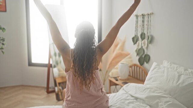 Young woman on bed stretching arms upward, bare back visible beside mirror and bedside table in bedroom; calm morning.