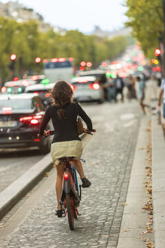 Woman rides a bicycle through busy Paris traffic on a dedicated cycle lane, surrounded by autumn trees, cars, pedestrians and colorful lights