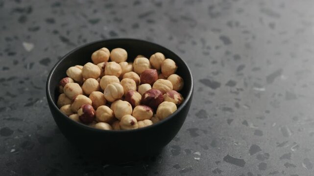 Slow motion handheld shot of roasted hazelnuts in black bowl on terrazzo countertop