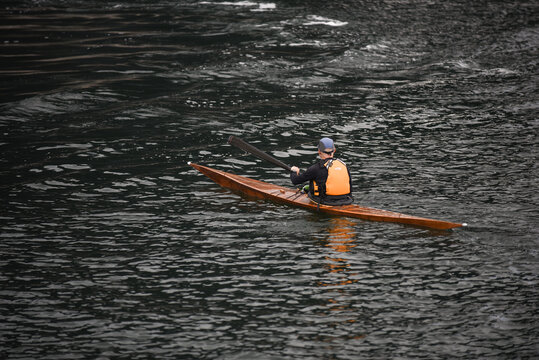 Aerial view of a person paddling in a wooden kayak with an orange life vest on the dark water of a canal in Copenhagen, Denmark.