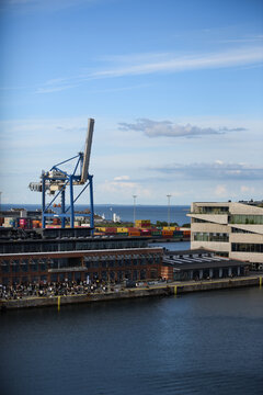 Aerial view of the industrial harbor area with a large blue crane, shipping containers, modern architecture, and people gathered along the waterfront in Copenhagen, Denmark.