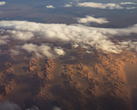 Aerial view of the vast Sahara Desert featuring rolling sand dunes, rugged terrain, and scattered clouds under soft light, Algeria.