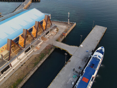 Aerial view of Waisai Harbour with a ferry docked at the concrete pier, modern terminal building with triangular roof structures, and calm waters in Raja Ampat, Papua Barat Daya, Indonesia.