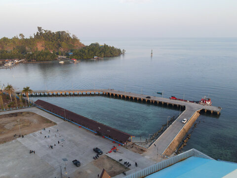 Aerial view of Waisai Harbour featuring a long concrete pier extending into the sea, with coastal area, lush hills, and vehicles in Raja Ampat, Papua Barat Daya, Indonesia.