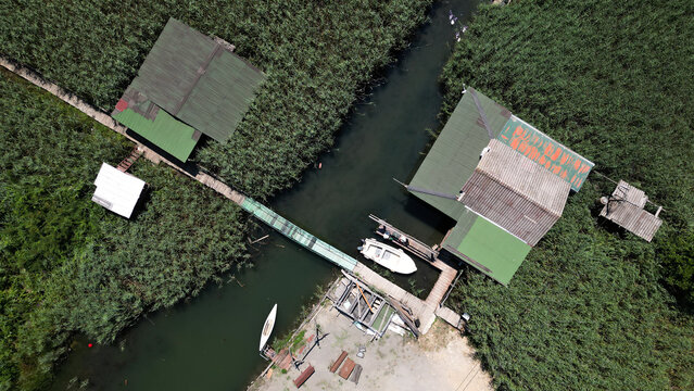 Aerial view of Ada Bojana featuring wooden huts with green roofs along a riverbank surrounded by dense reeds and a small boat moored at a wooden pier in Ulcinj Municipality, Montenegro.