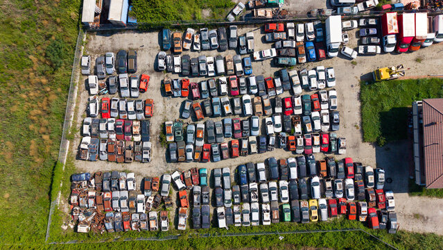 Aerial view of a large car scrapyard filled with rows of parked vehicles in various colors, surrounded by a fence and grassy fields in San Giuliano Milanese, Lombardy, Italy.