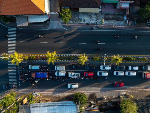 Aerial view of a busy road with cars and motorcycles waiting at a crosswalk, lined with palm trees in Kecamatan Denpasar Selatan, Bali, Indonesia.