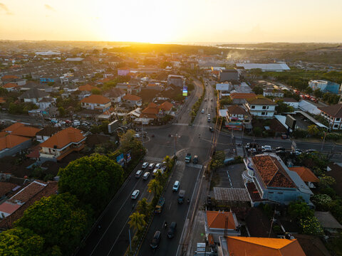 Aerial view of a busy road intersection at sunset, with vehicles, traditional buildings with orange roofs, lush trees, and golden light in Kecamatan Denpasar Selatan, Bali, Indonesia.