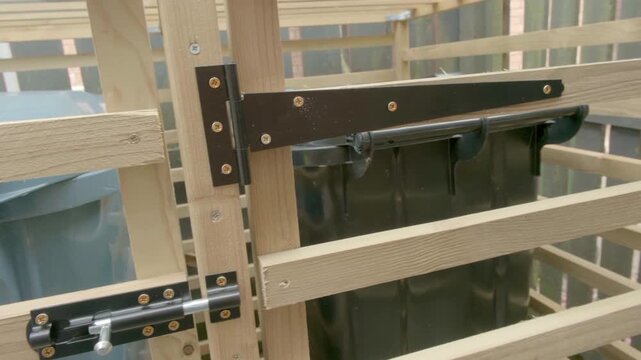 Wooden gate to wooden constructed bin hide with metal latch and bolt. Latch secures structure for safety. No people appear in footage. Ideal for animal enclosures or storage waste bins.