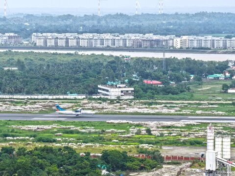 Aerial view of a passenger airplane landing on the runway at Cox's Bazar Airport surrounded by lush greenery and buildings in Cox's Bazar, Chittagong Division, Bangladesh.