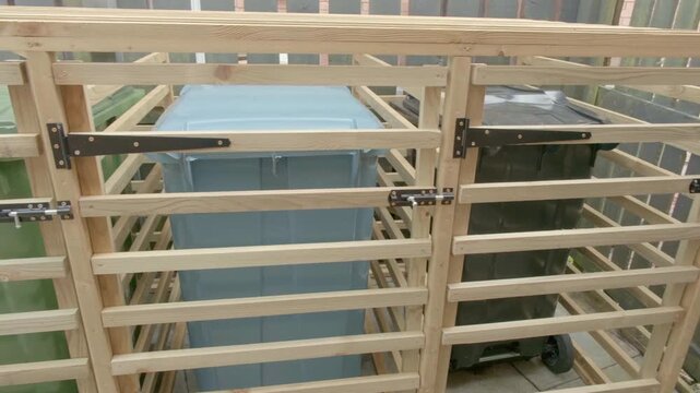 Wooden bin hide with steel hinges and brass screws. Green and blue bins secured within protective frame. Simple design hides waste from curious eyes. Ideal for garden or backyard settings