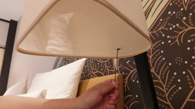 Hand pulling the chain cord switch of a modern bedside lamp in a cozy hotel bedroom.