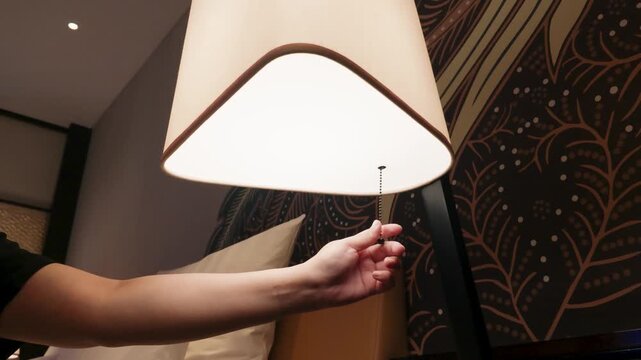 Person hand pulling switch to turn on bedside lamp in cozy bedroom.