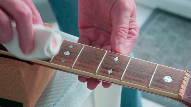 Hands gently wipe guitar fretboard with white cloth polishing the fret board. Fingers move carefully over wood grain and metal frets. Guitar rests on soft, textured surface. Calm focus evident as care