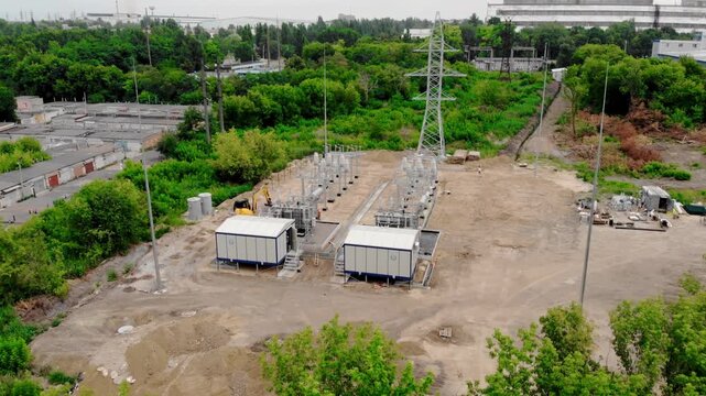 Aerial drone view of an electrical substation under construction in Sumy, Ukraine. High-voltage transformers, switchgear, transmission equipment, utility buildings, workers, and a yellow excavator