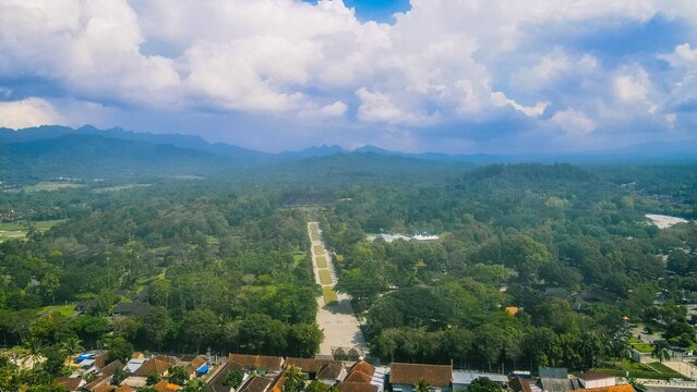Aerial view of the historic Borobudur temple complex surrounded by lush tropical greenery and distant mountains under a cloudy blue sky in Borobudur, Central Java, Indonesia.