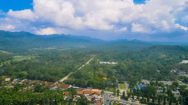 Aerial view of the historic Borobudur temple complex surrounded by lush tropical forests, mountains, and local village rooftops under a bright blue sky in Borobudur, Central Java, Indonesia.