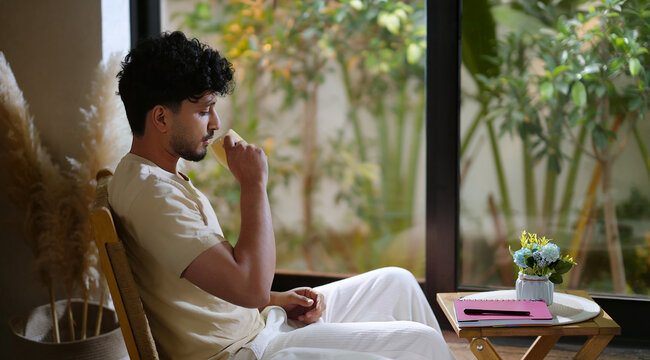 Indian calm young adult gen z relax man sitting on chair hold tea mug drink coffee enjoy day room male boy spend time take casual job break home house place