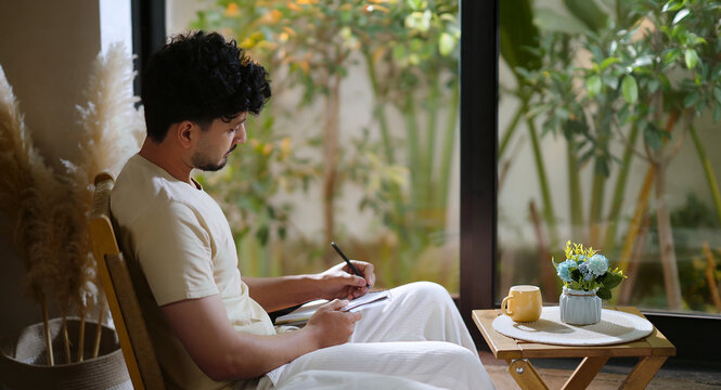 Indian calm young adult gen z man sitting on chair enjoy day resting indoor room. Male boy hold pen writing book new work notes plan ideas spend time take casual job break home house place
