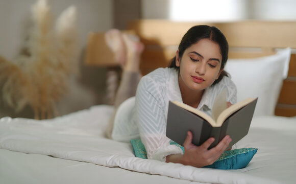 Indian young adult gen z relax calm woman lying on cozy bed read new novel book story indoor room resting girl spend free time do self care enjoy day take break from job work home place