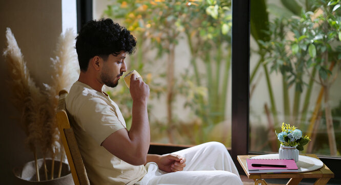 Indian calm young adult gen z relax man sitting on chair hold tea mug drink coffee enjoy day room male boy spend time take casual job break home house place