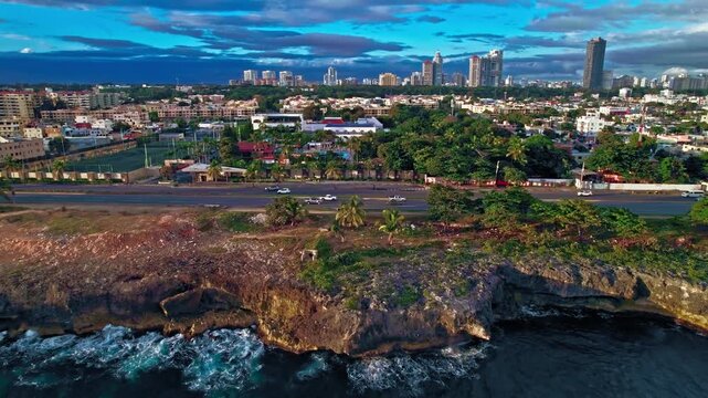 Daytime aerial drone shot moving forward over Malecon coastal road to reveal Casa de Espana social club pools, sports courts, and parking lot in Santo Domingo, Dominican Republic