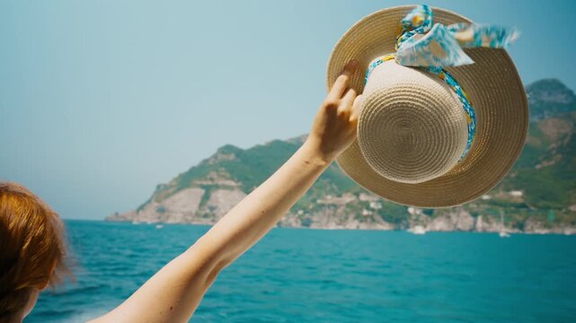 Arm lifting a straw hat as red hair blows loose in Amalfi coast