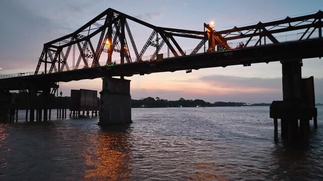 Cinematic view of historic Long Bien Bridge spanning the Red River at dusk, with glowing lights, silhouetted steel trusses, and reflections on the water.