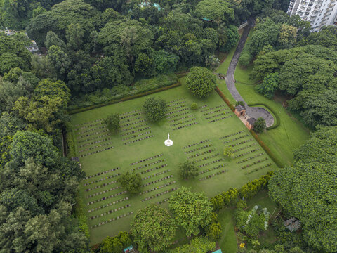 Aerial view of the Chittagong War Cemetery, featuring rows of gravestones, a central memorial, lush green grass, and surrounding dense trees in Chittagong, Chittagong Division, Bangladesh.